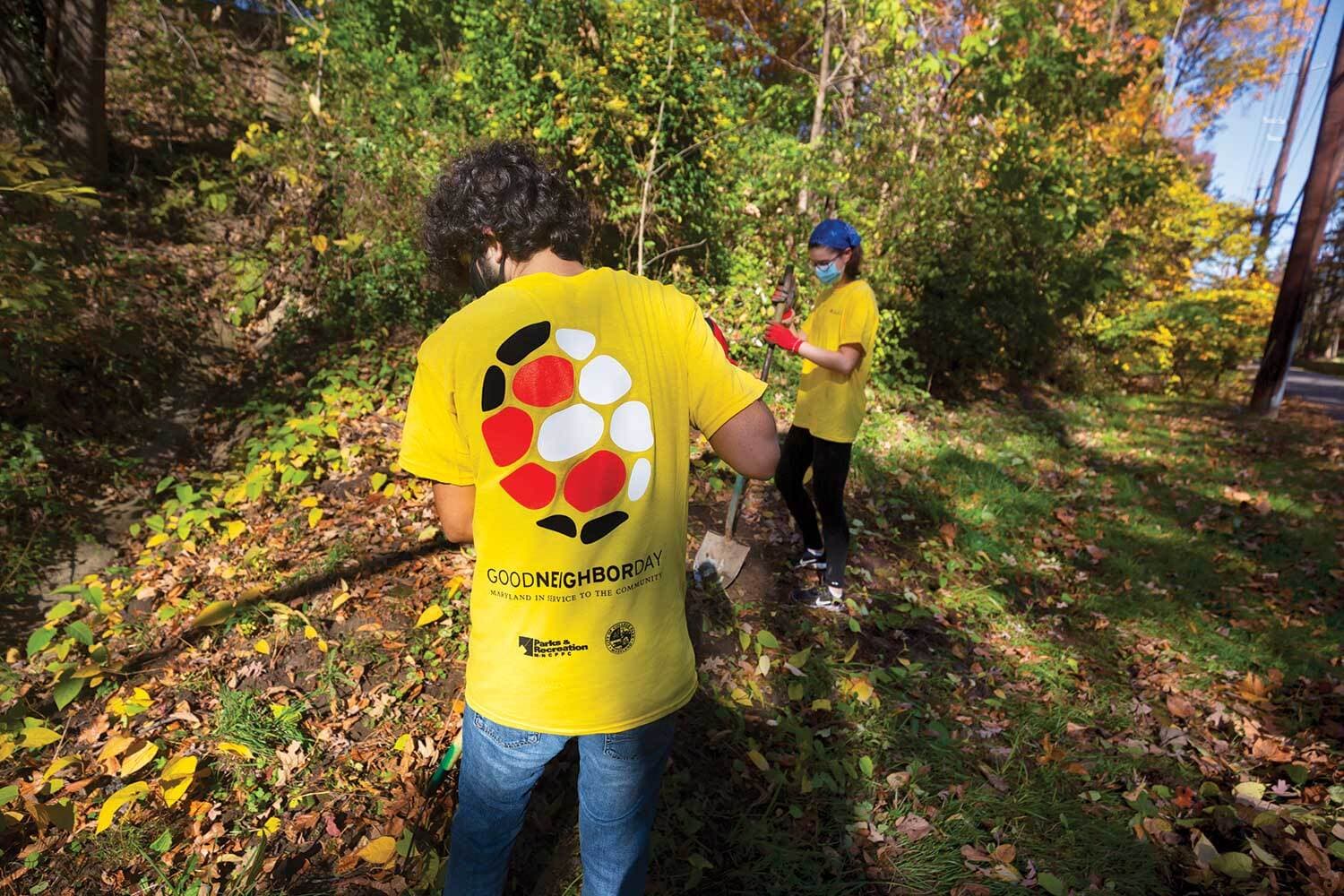 Two students wearing yellow shirts with a terrapin shell and Good Neighbor Day emblazoned on the back rake leaves in a wooded area.