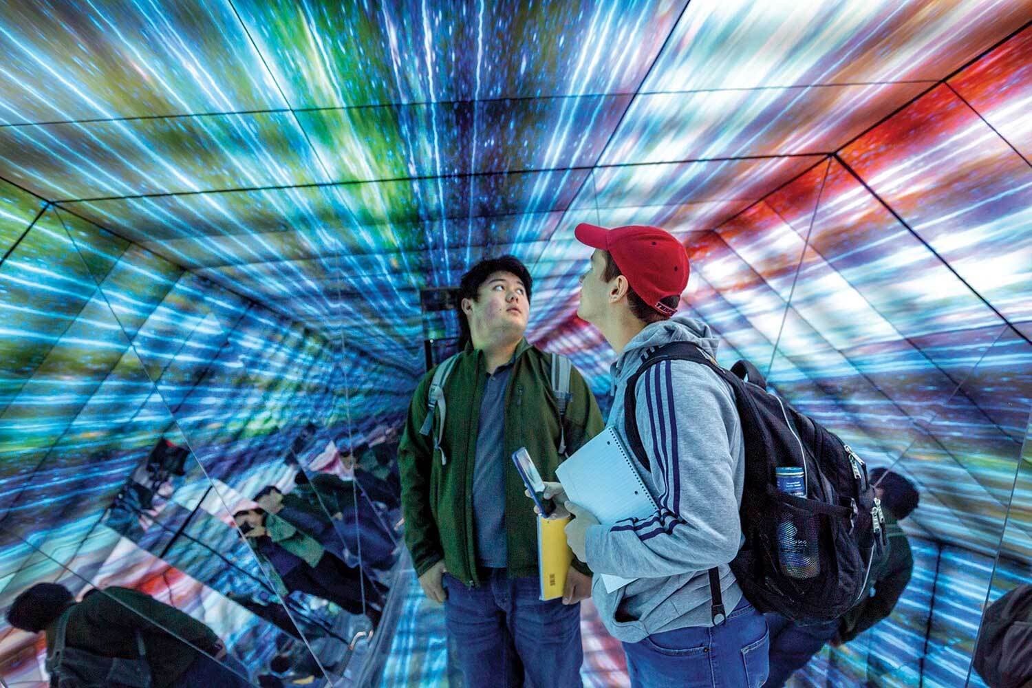 Two students stand amazed and confused in a hexagonal mirror-walled room with colored lines radiating from a single point that look like a science fiction warp drive but in color.