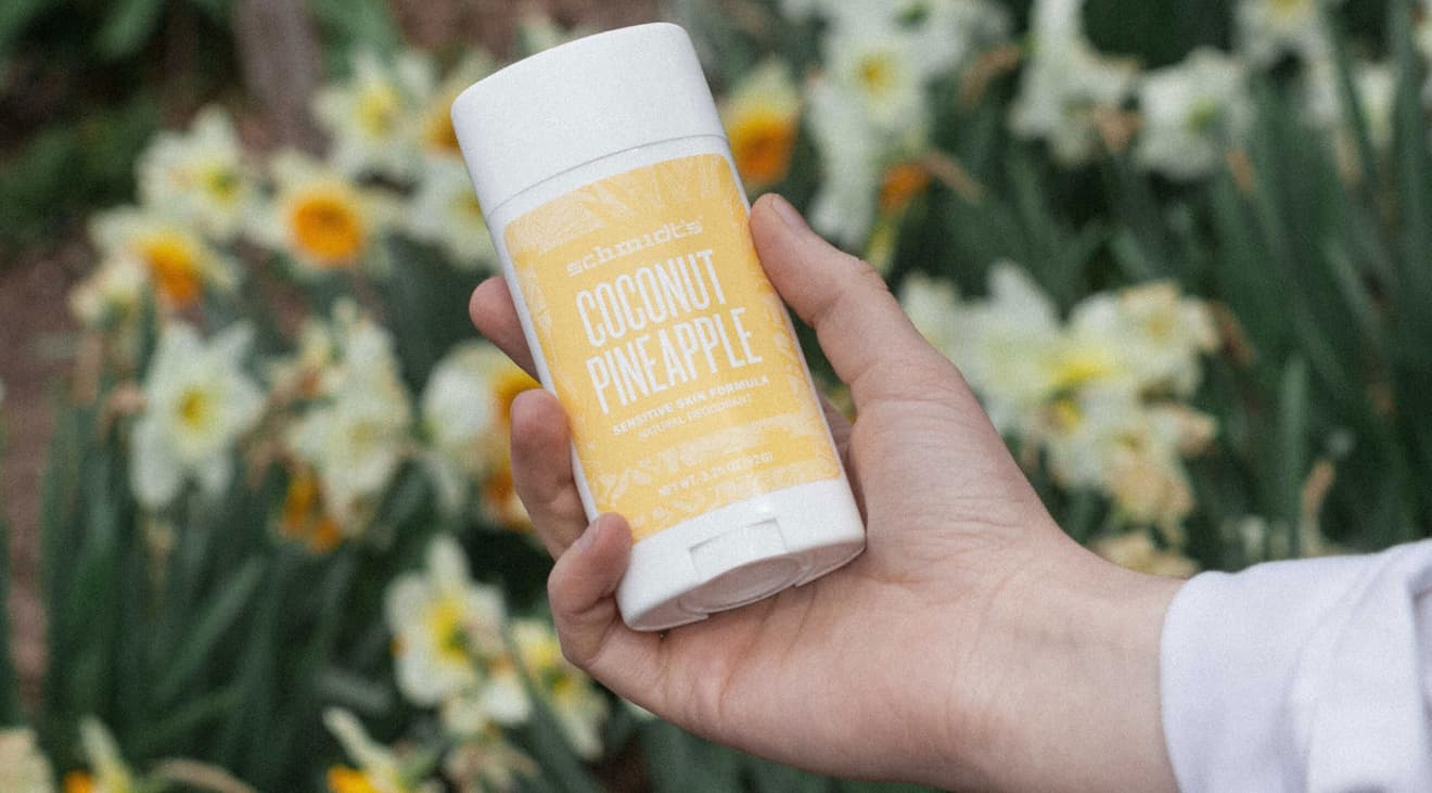 A hand holding a coconut pineapple scented stick of deodorant in front of flowers.