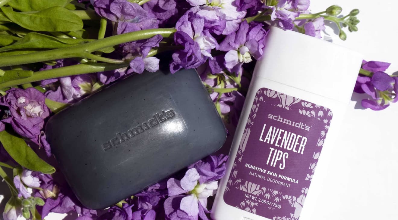 A charcoal bar of soap next to a lavender-scented stick of deodorant laid on top of purple flowers.