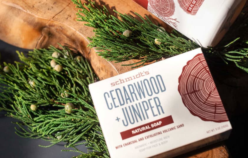 Boxes of cedarwood+juniper scented bars of soap sit on top of a piece of wood with some pine needles intertwined.