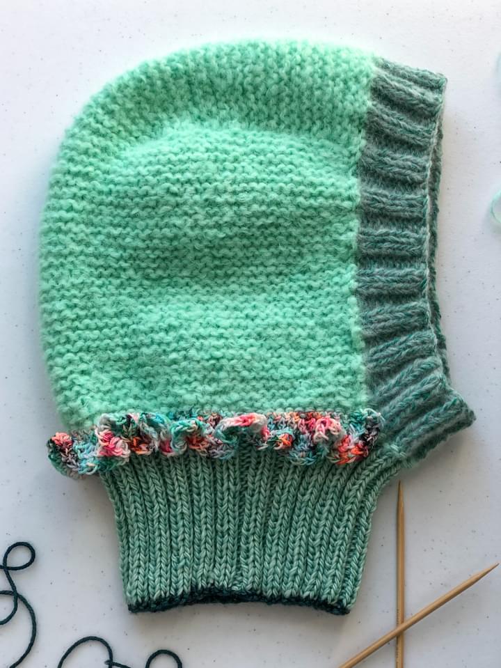 A knitted green balaclava hat with a multicolor ruffle laying flat on a table by some knitting needles