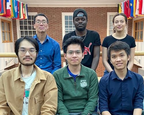 A group of six diverse students posing for a photo in a room adorned with international flags, representing a collaborative research environment under the guidance of Professor Peter Chin for a DARPA Cybersecurity Research Grant project.