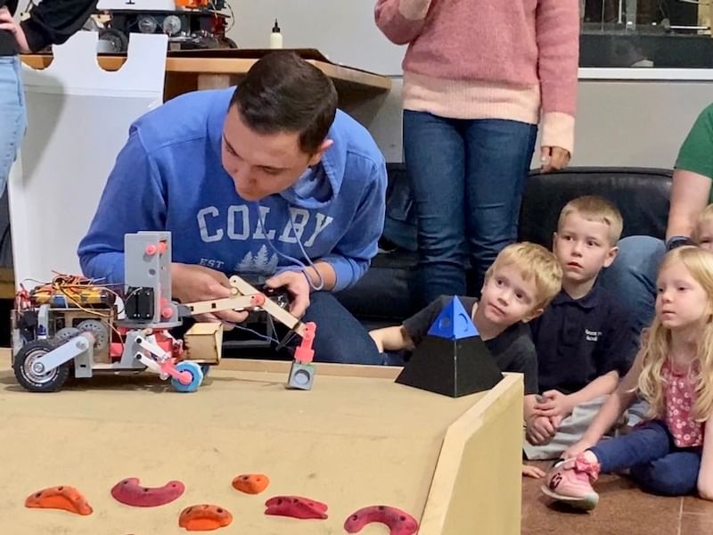 Student wearing a blue COLBY sweatshirt, intently adjusting a small robotic rover with a mechanical arm, in front of a captivated audience of young children.