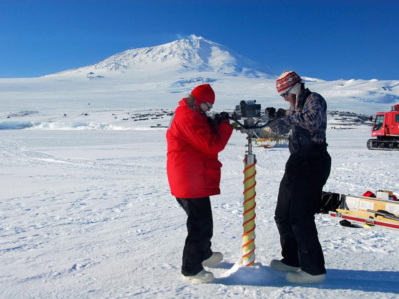 Two people stand on either side of a drill in on a large sheet of ice while they perform Ice Core Drilling