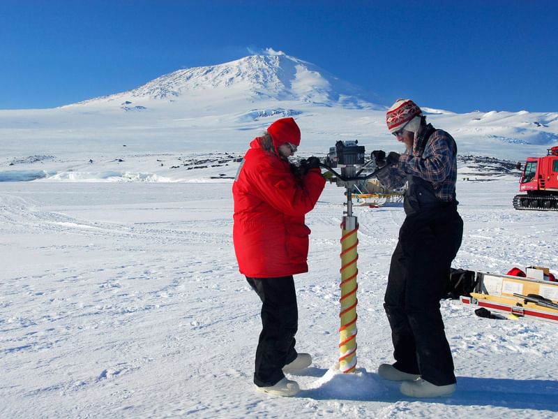 Two researchers in cold-weather gear are extracting an ice core sample in a snowy Antarctic landscape, with a mountain in the background and a red vehicle to the side.