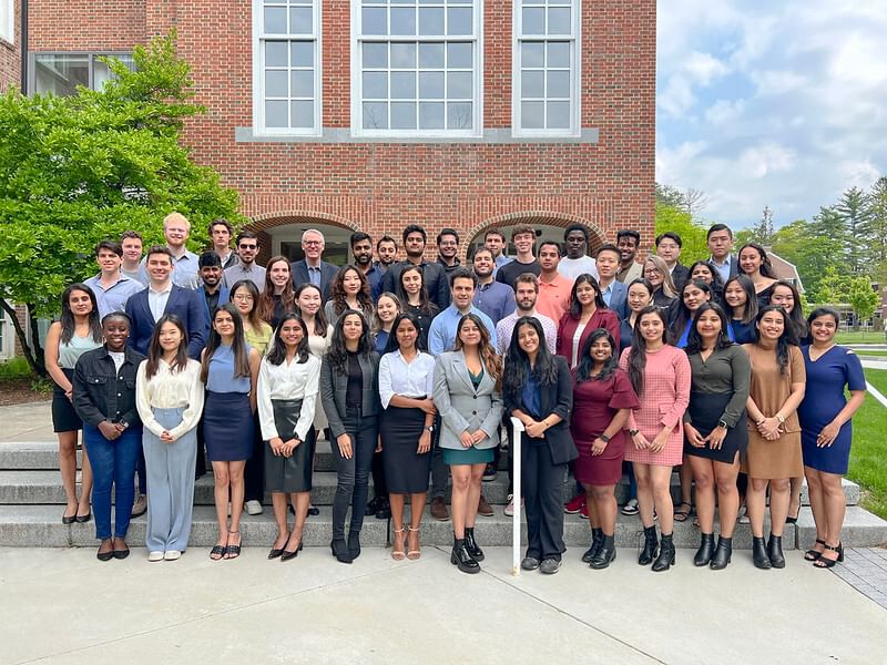 A diverse group of students, dressed in business attire, is posing for a photo in front of a brick building, representing a cohort from an Dartmouth MEM focused on blending technical and business education.