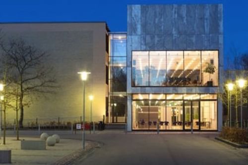 The entrance to a building at the Technical University of Denmark – DTU is visible at dusk, showcasing modern architecture with large glass windows that reveal the illuminated interior. The pathway to the well-lit entrance is lined with trees and spherical bollards.
