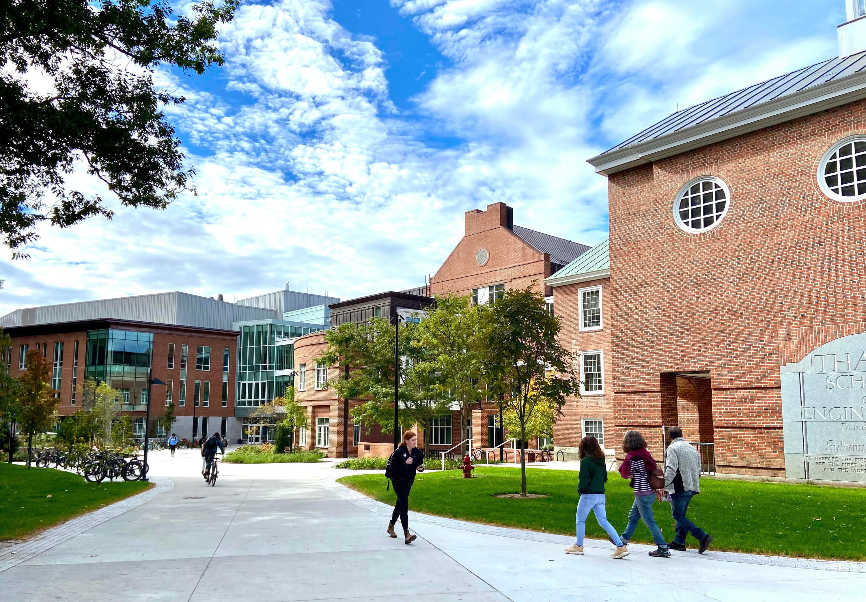 Under a blue sky, students walk along a pathway next to brick buildings on the West End of Dartmouth's campus.