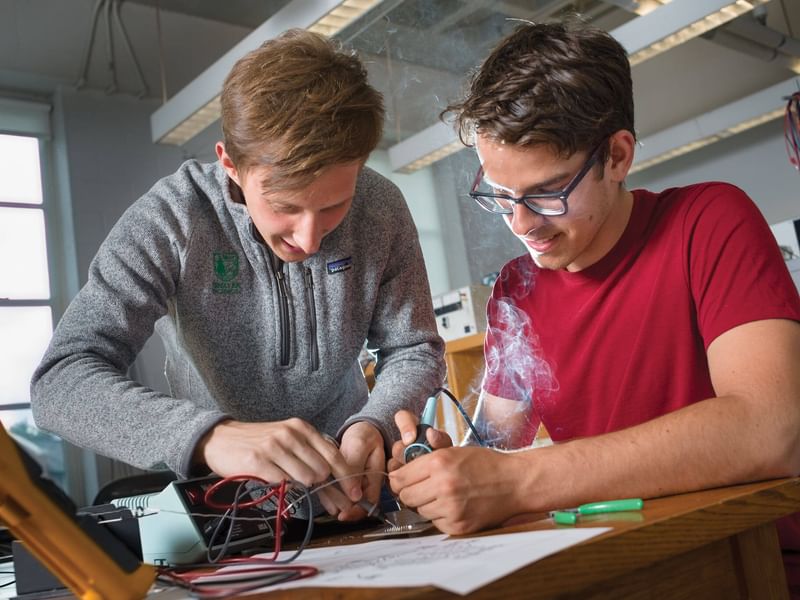 Two students are engaged in assembling electronic circuits, with one soldering parts as smoke rises. They are focused on their task in a laboratory setting. One has a grey shift and the other is red.