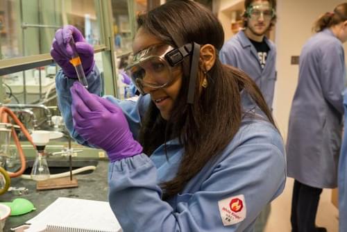 A student wearing protective lab gear looks closely at liquid in a test tube at a lab bench.