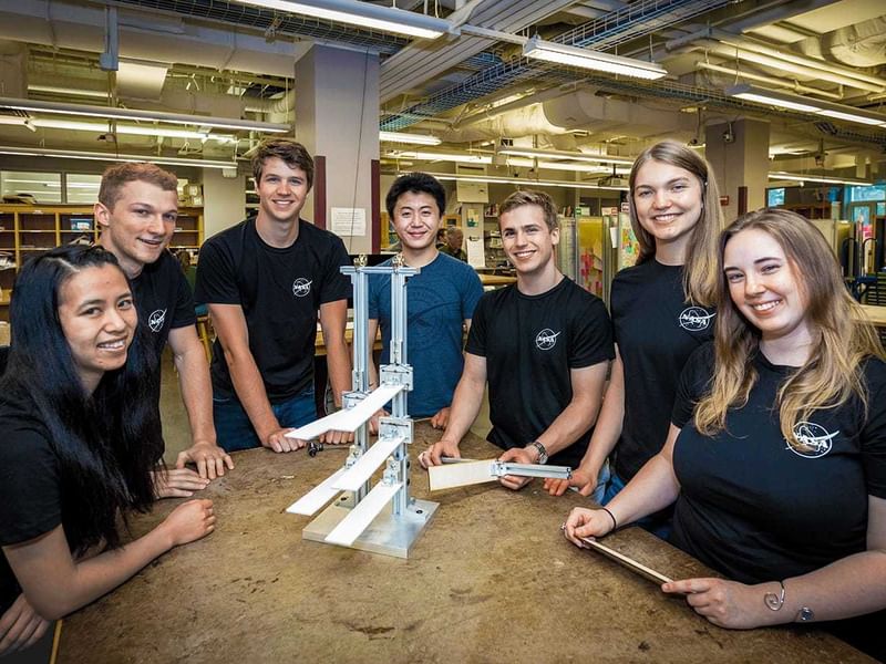 A team of seven engineering students stands proudly around a mechanical prototype at the Cook Engineering Design Center, exemplifying the hands-on, collaborative projects designed to address real-world challenges such as space agriculture for NASA. They are dressed in matching black team shirts