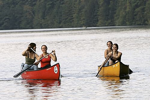 Four people in two canoes are paddling on calm water. The first canoe is red with two people wearing life jackets, one paddling on the starboard side. The second canoe is yellow with two people; the person in the stern is paddling. Both canoes have registration numbers on the bow. Trees reflecting on the water.