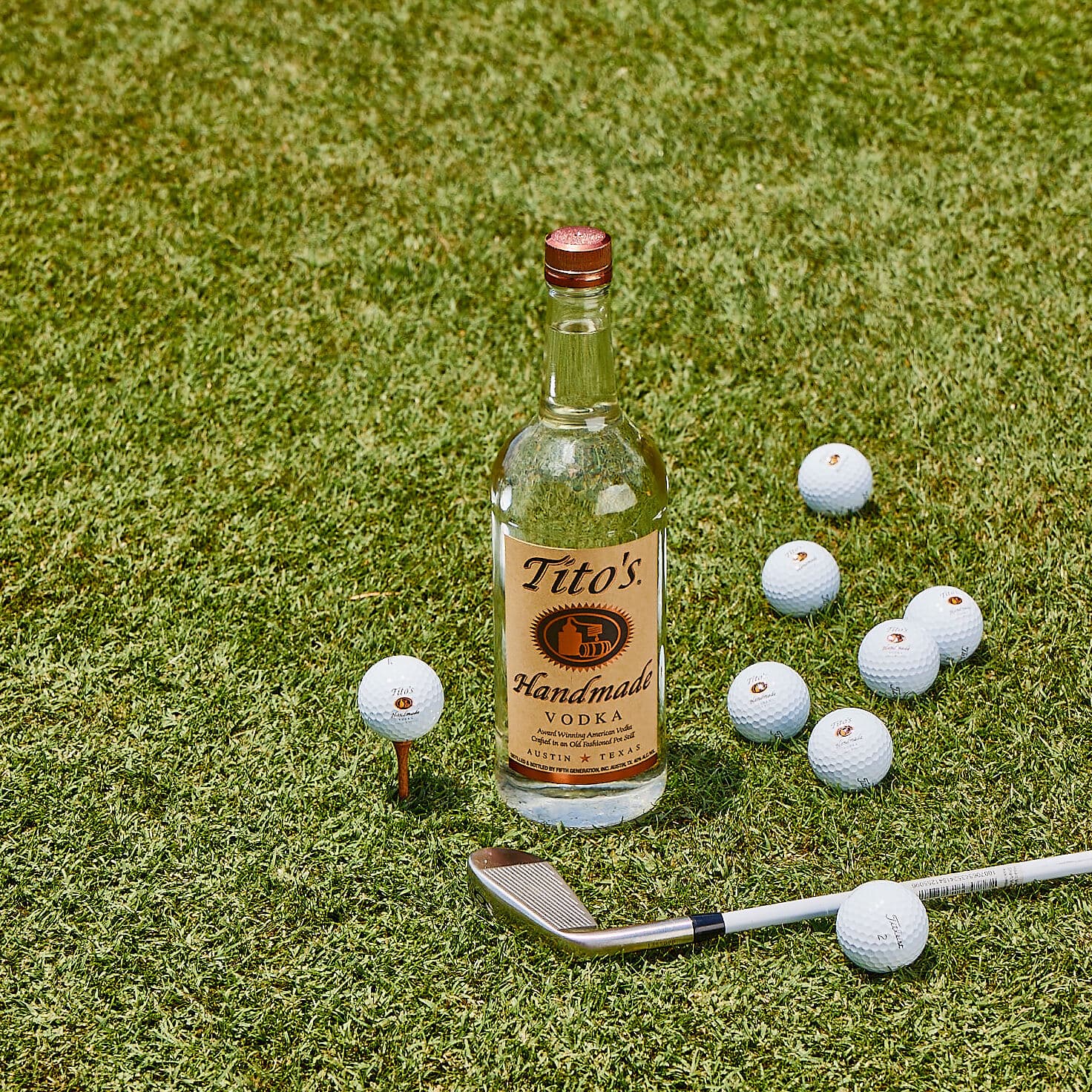 Tito's Handmade Vodka bottle on putting green with Tito's golf balls and silver putter