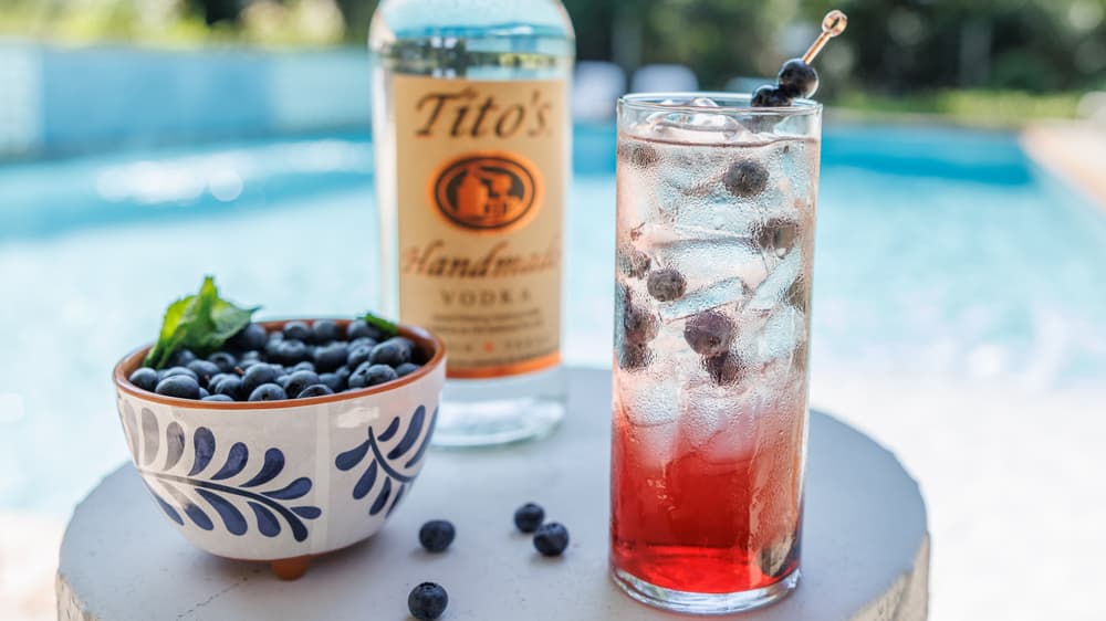 Tito's Berry Sparkler cocktail, a bottle of Tito's Handmade vodka, and a bowl of blueberries in front of a pool