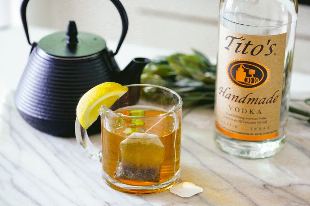 Tito's Toddy in a glass mug with a lemon garnish, a tea kettle and bottle of Tito's Handmade Vodka in the background