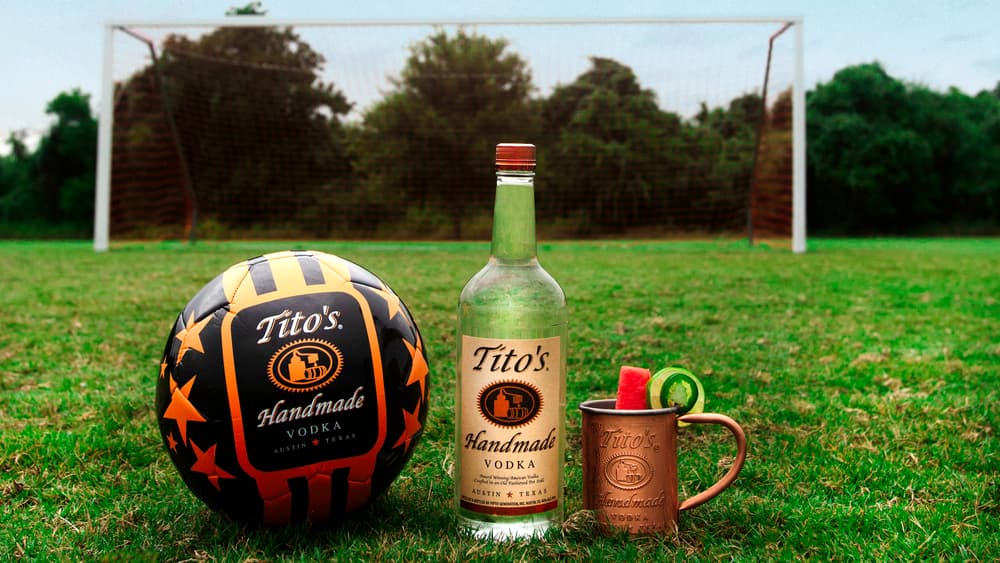 Tito's Vodka bottle with a Tito's branded soccer ball and a cocktail in a copper mug