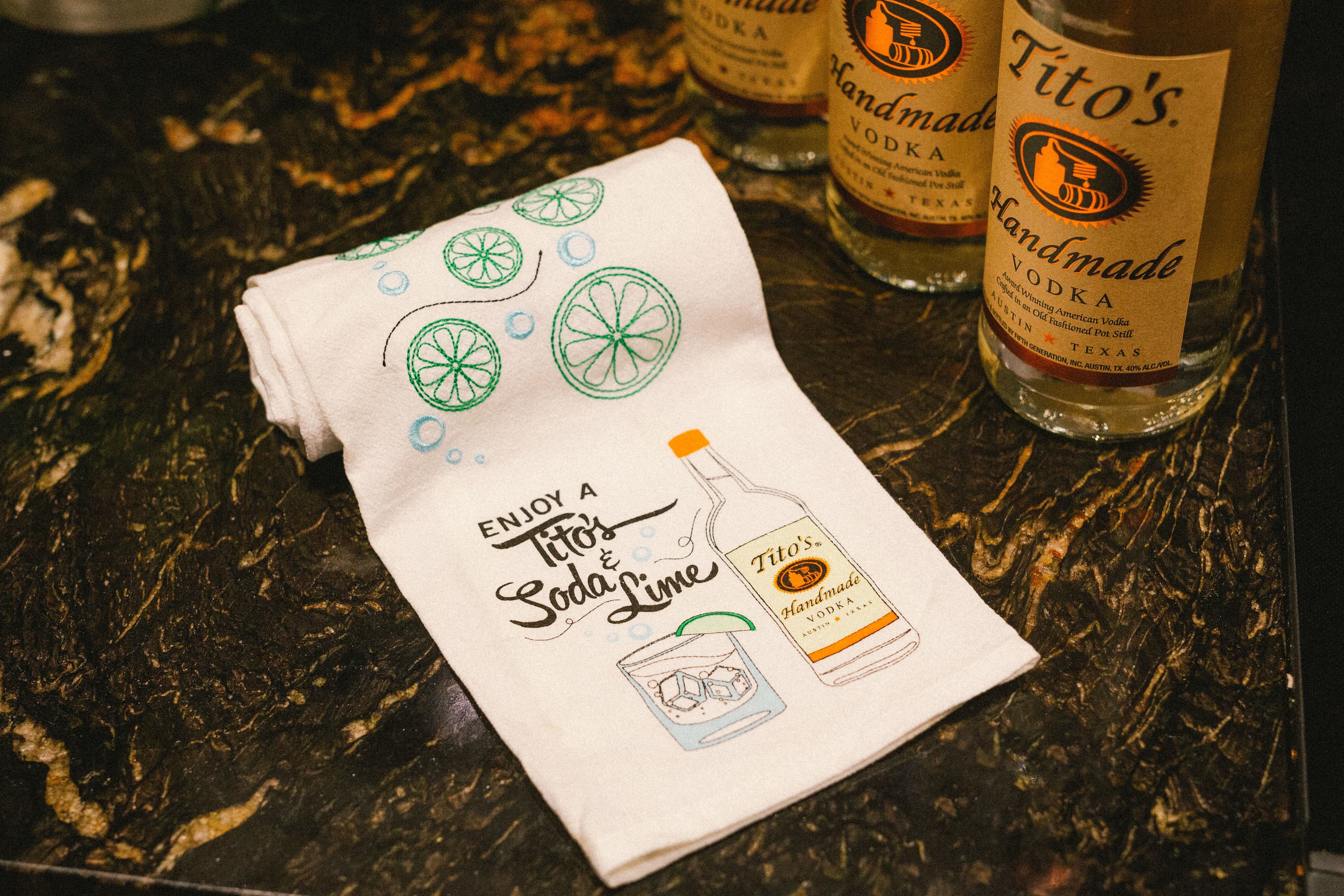 White tea towel with printed Enjoy a Tito’s Soda & Lime design and embroidered limes next to bottles of Tito's Vodka