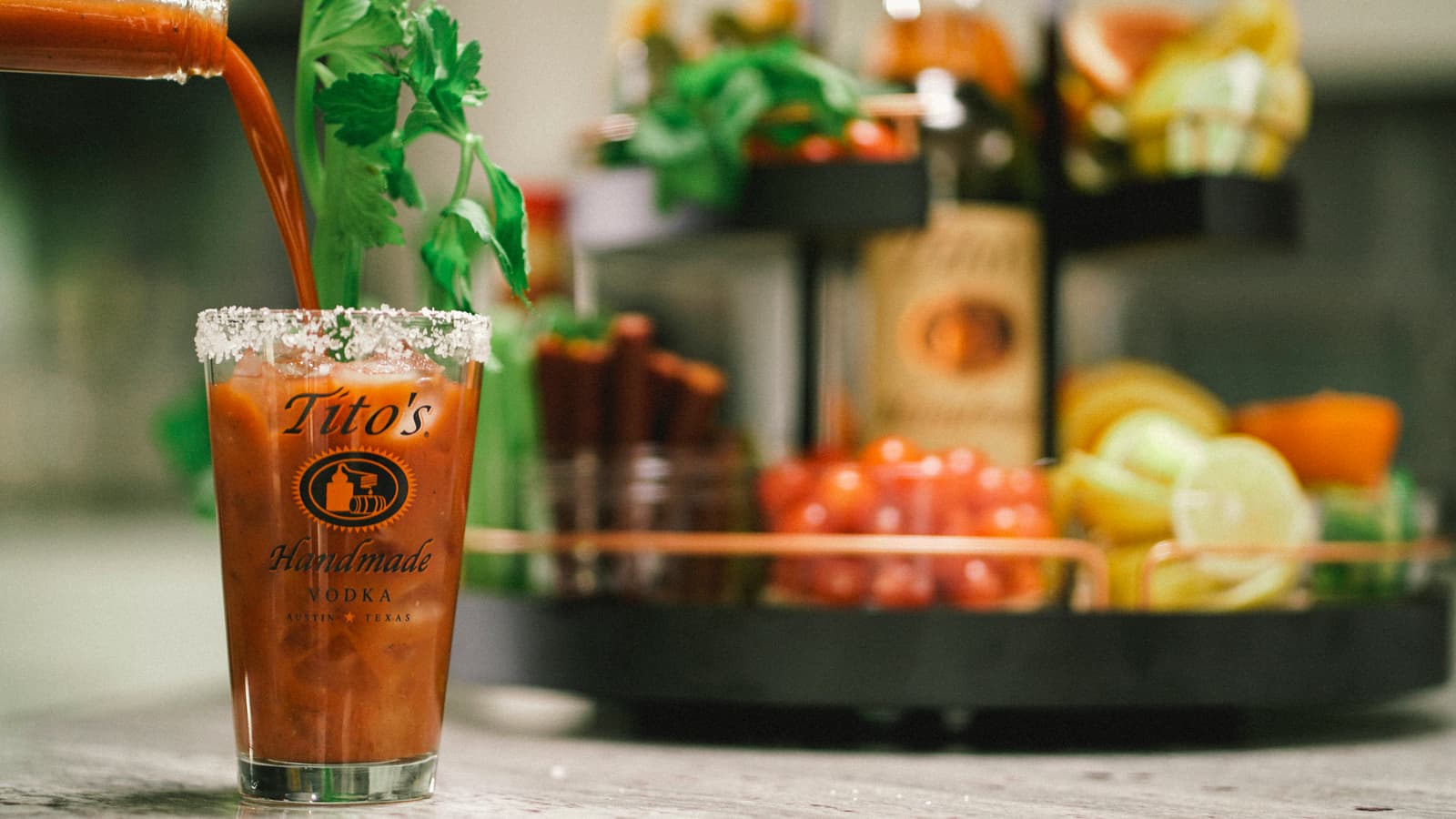 Bloody Mary being poured into Tito's Pint Glass with tray of garnishes in the background
