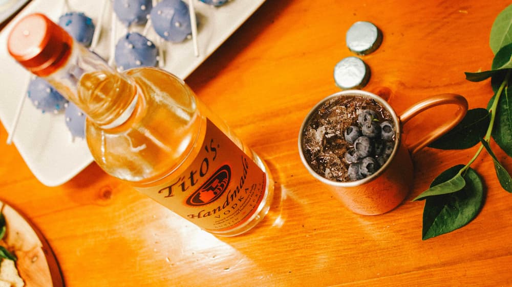 Tito's Vodka bottle next to a Blueberry Mule in a copper Mug garnished with blueberries