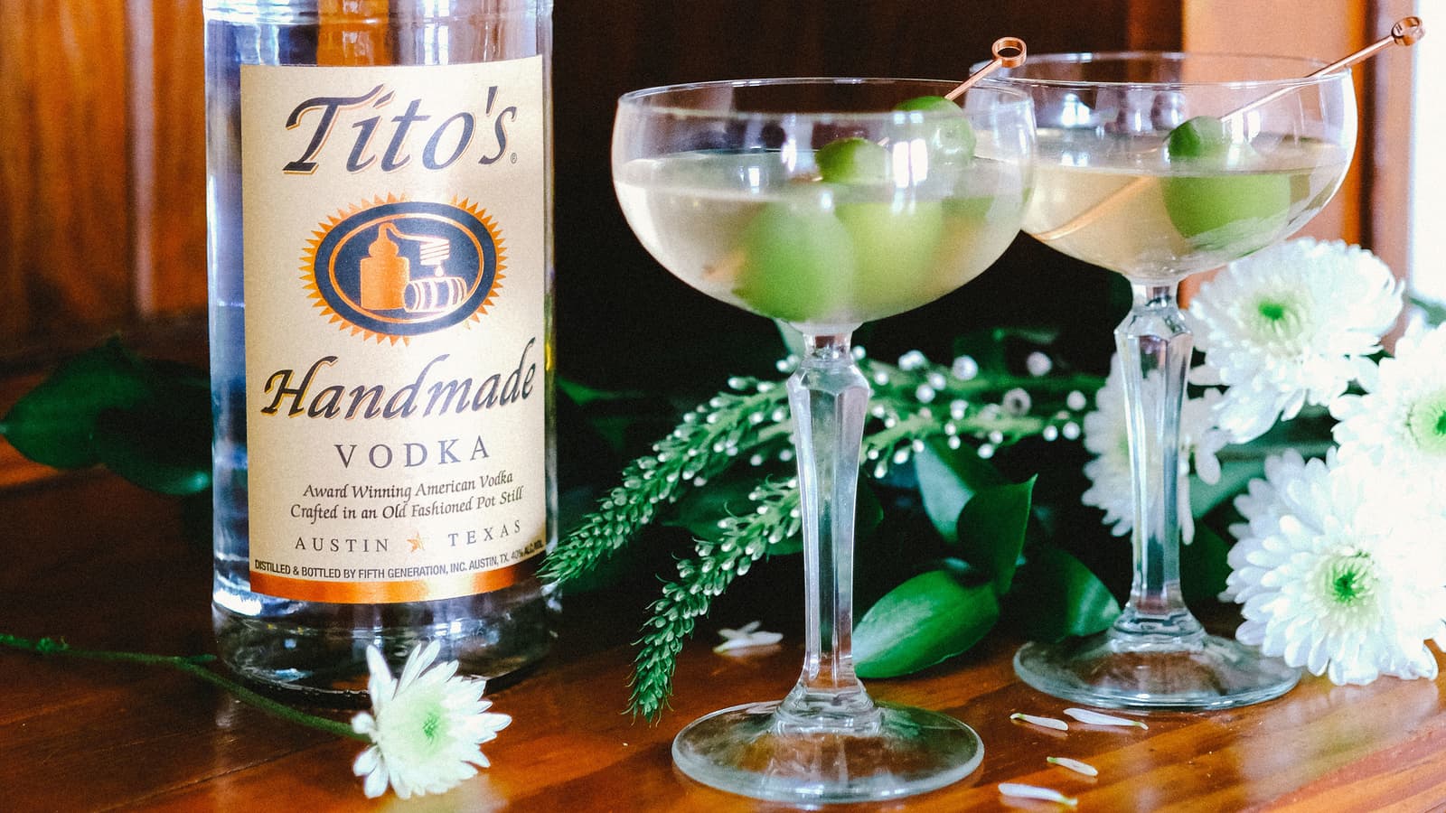 Two Olive You So Much cocktails on a table with white flowers and a Tito's Vodka bottle