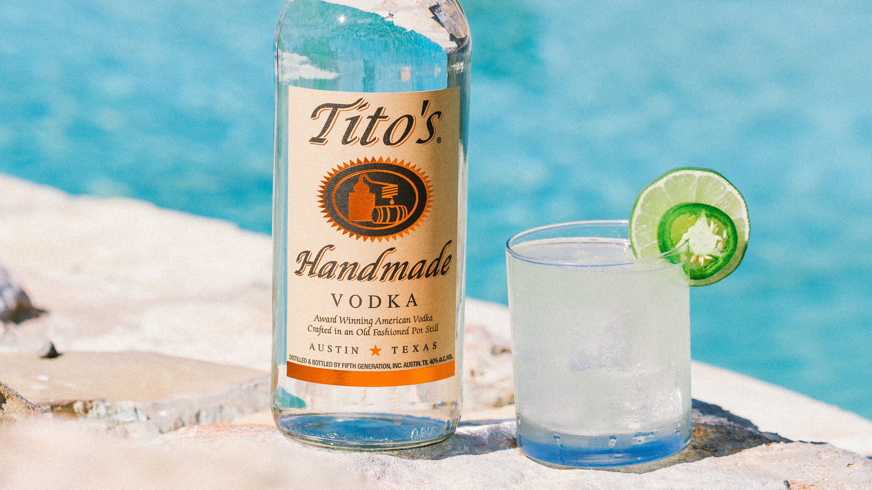 Summer Heat cocktail garnished with a lime and jalapeno next to a Tito's Vodka bottle in front of a swimming pool
