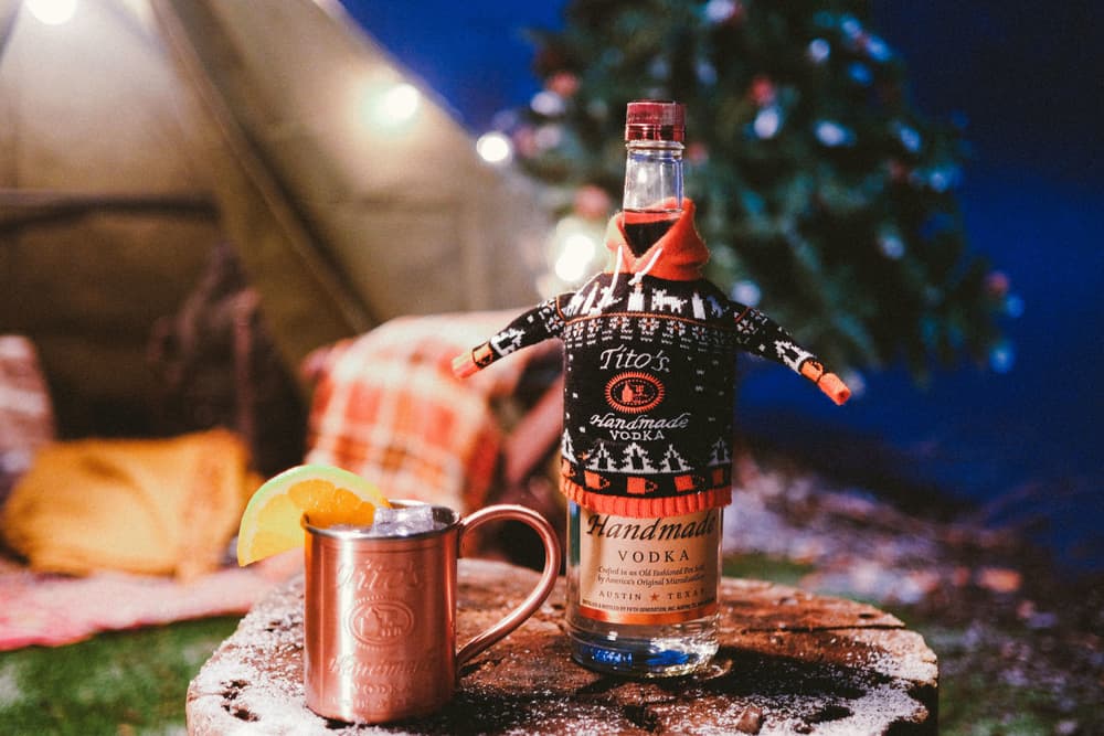 Copper mug garnished with an orange and Tito's Vodka bottle in a holiday sweater on a tree stump