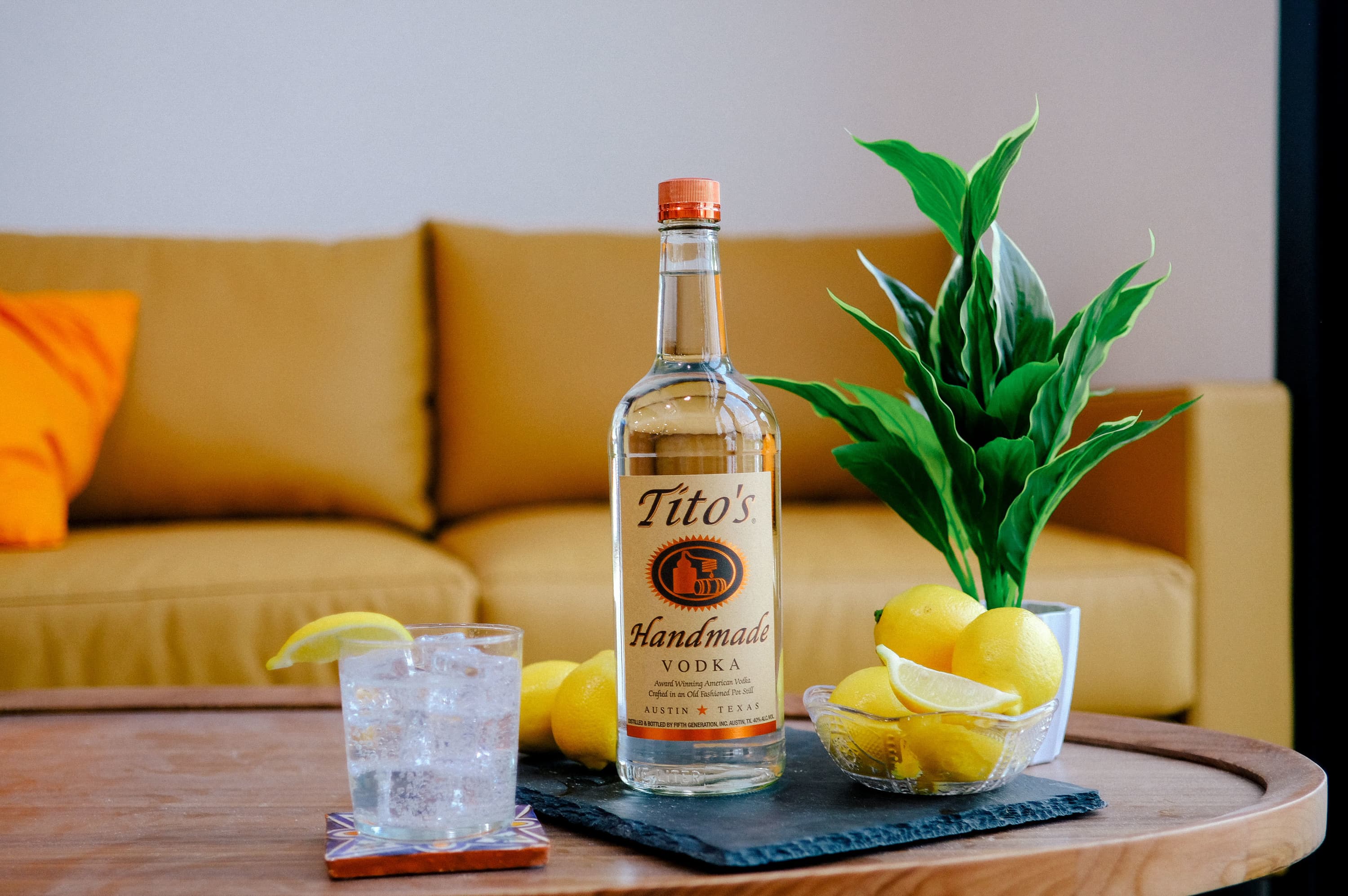 A bottle of Tito's Handmade Vodka on a table with a bowl of lemons and a Tito's Water & Lemon cocktail