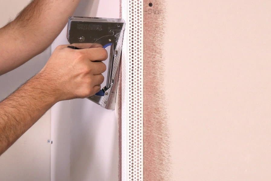 An image of a person’s hands holding a staple gun and applying vinyl corner beads to a wall.