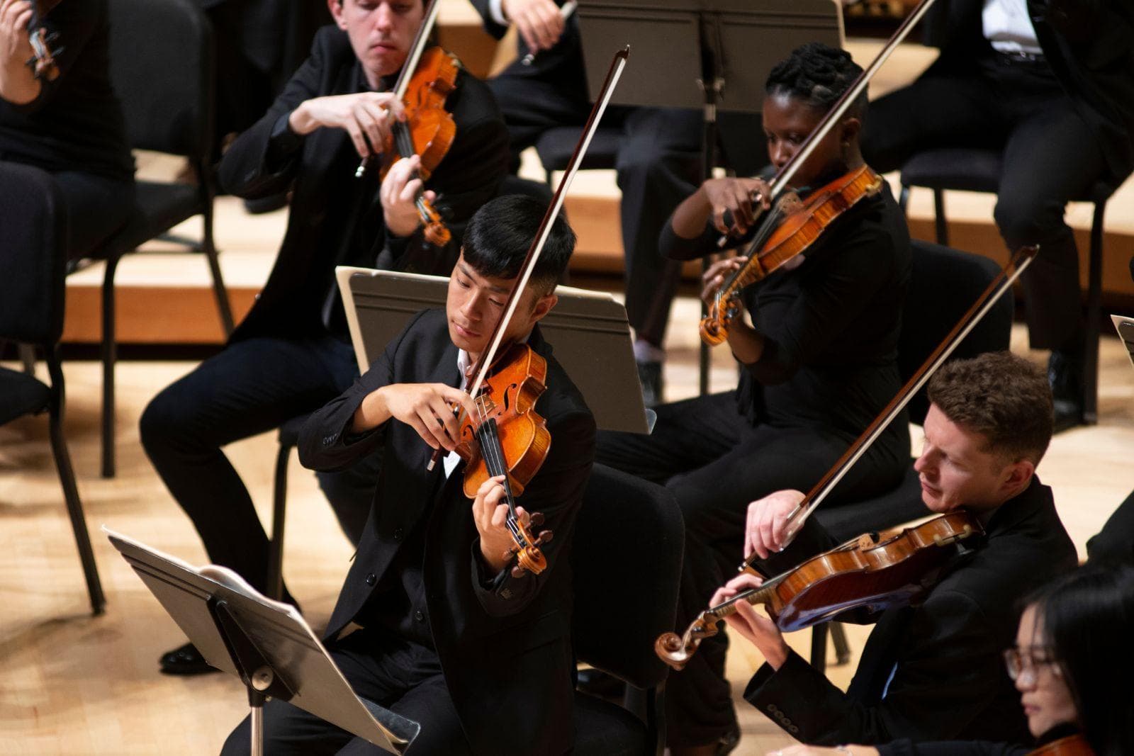 Students play violins in an orchestra on stage.