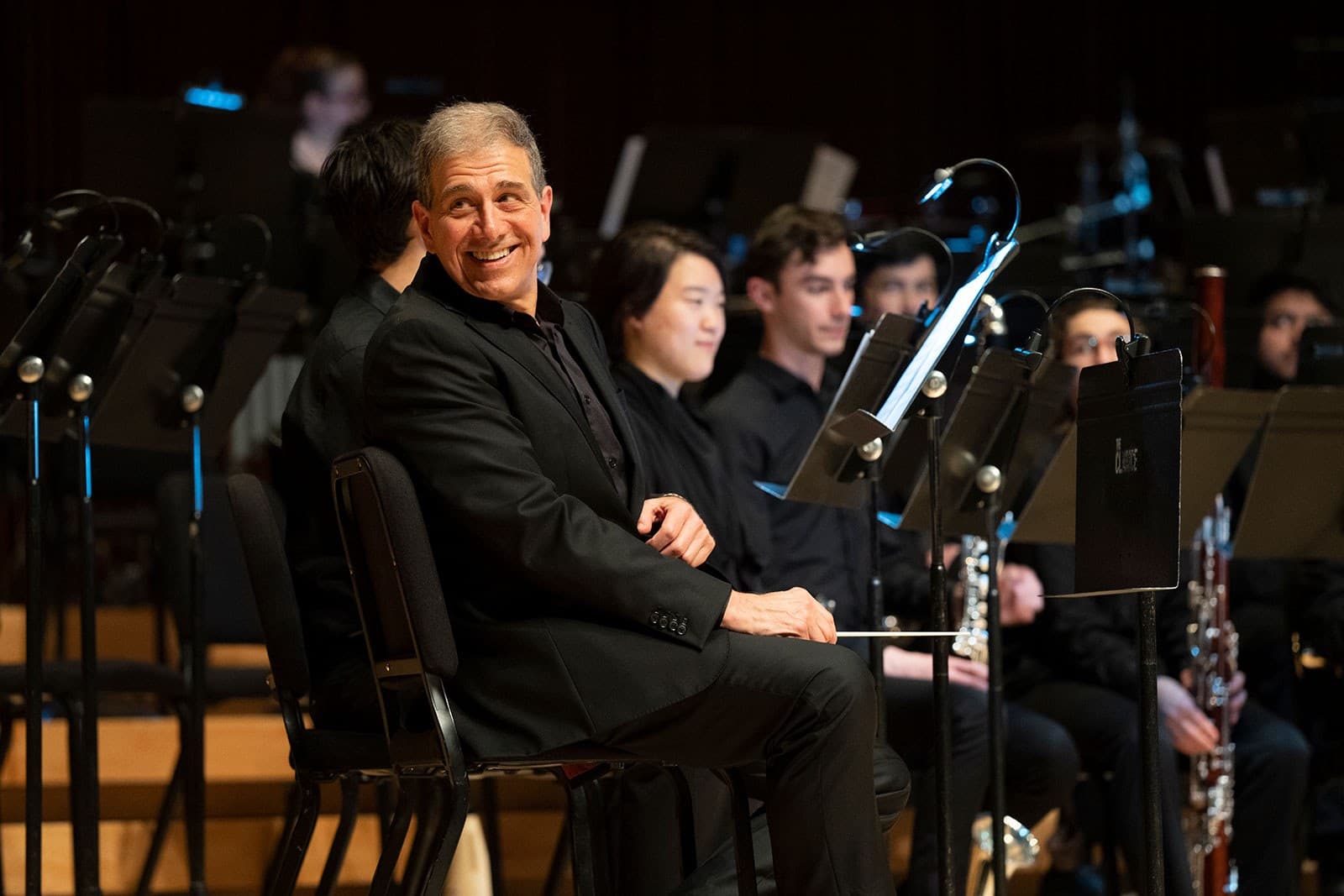 Man sitting on stage among an orchestra smiles off in the distance.