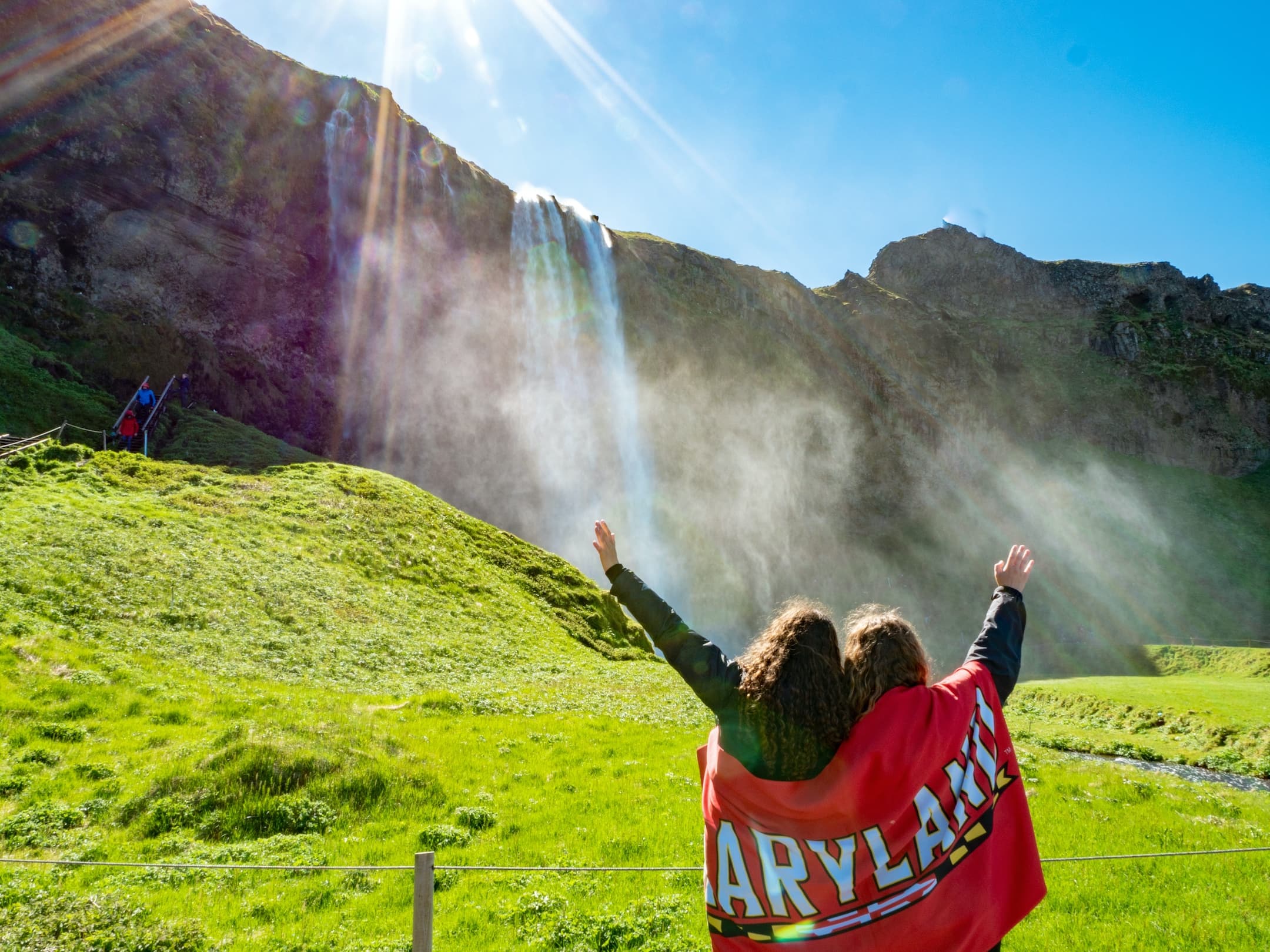 Two students stand observing a large waterfall with their backs to the camera. A UMD flag is draped across their shoulders and they each raise an arm to the sky.