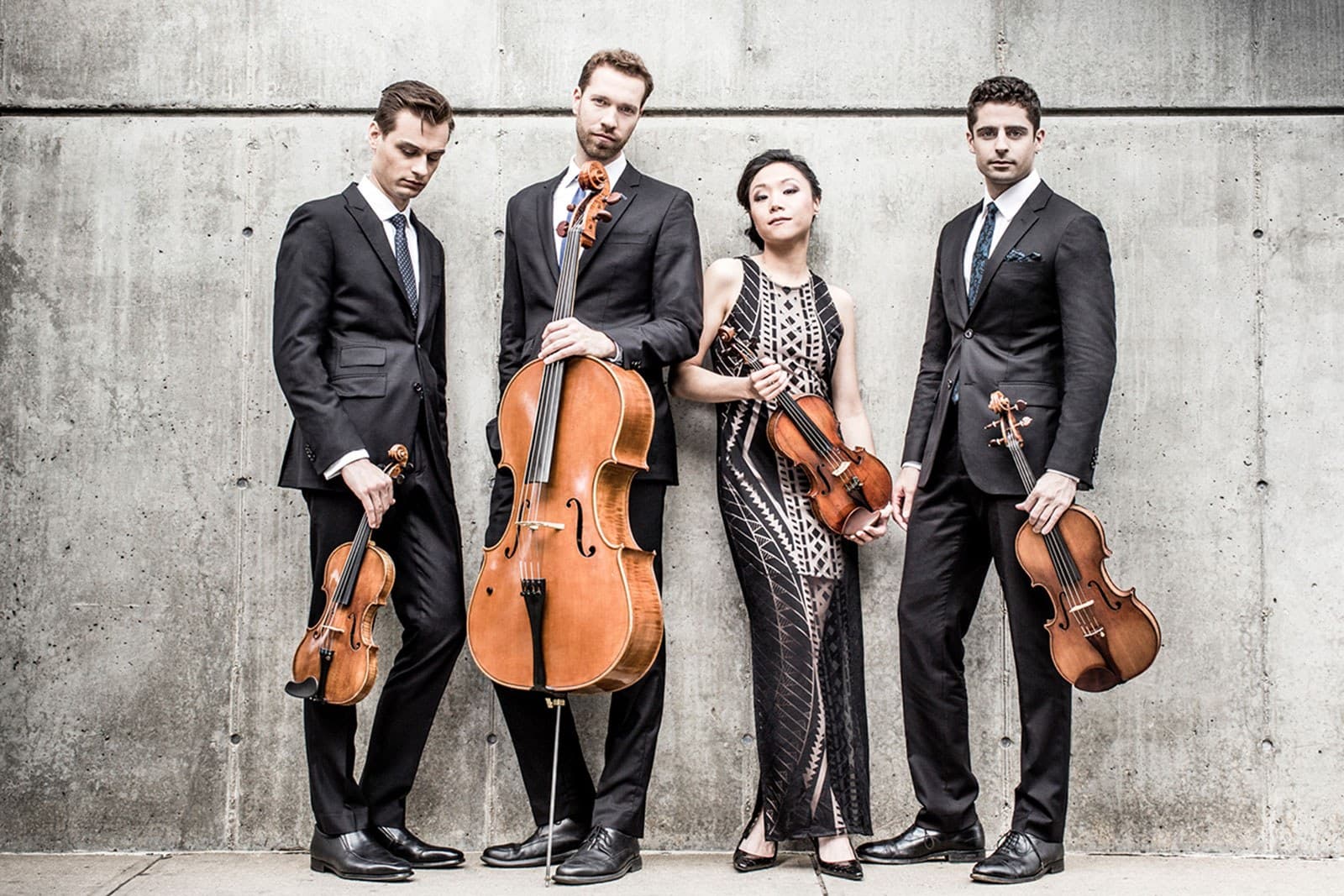 Three men and a women pose against a wall holding violins and a cello.