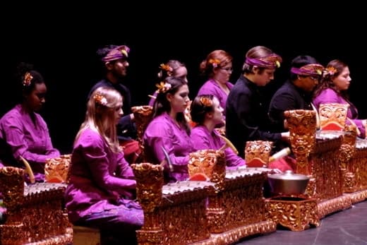 Students wearing traditional clothing play Balinese instruments on stage.