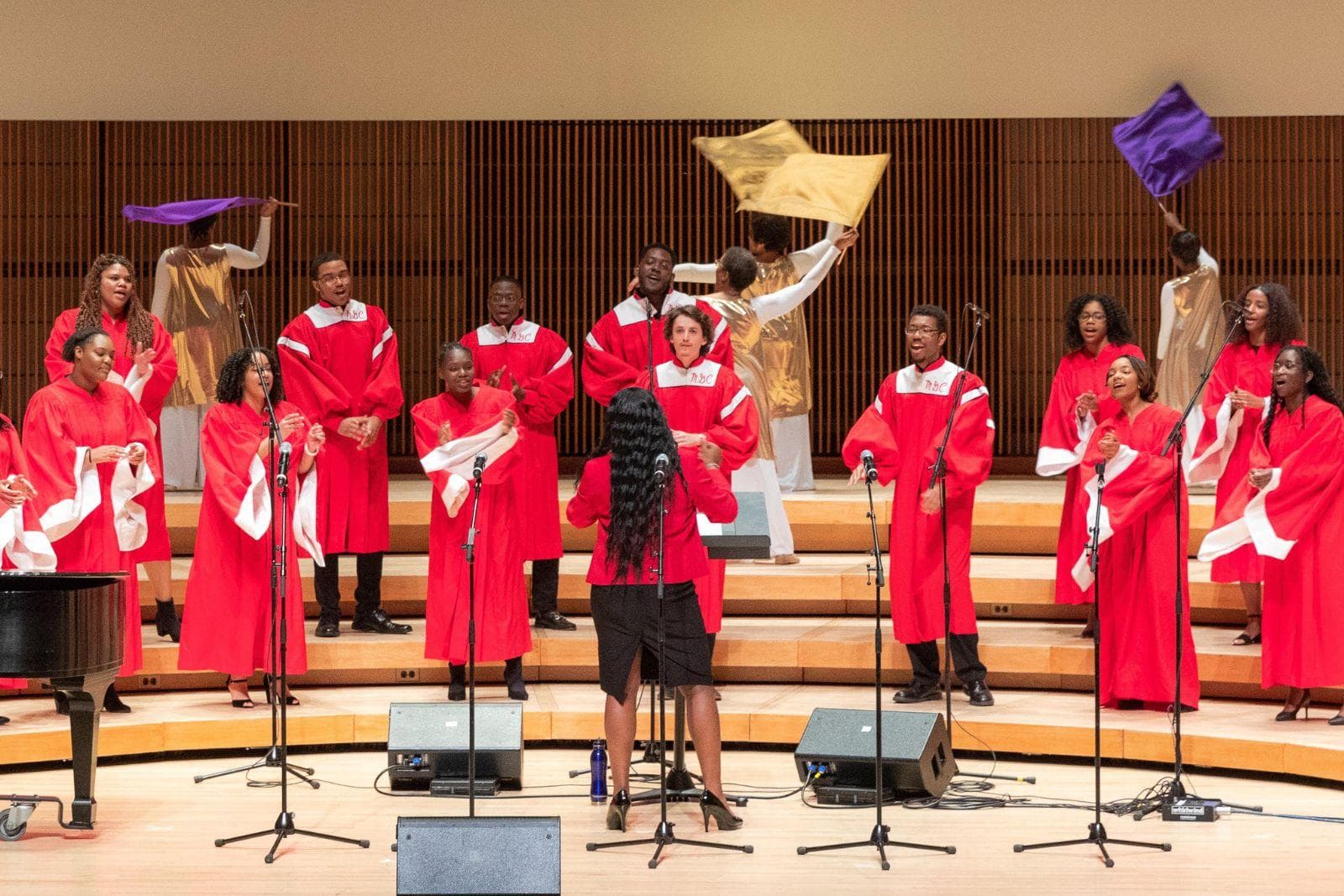 Gospel choir members wearing red robes perform in a concert hall.