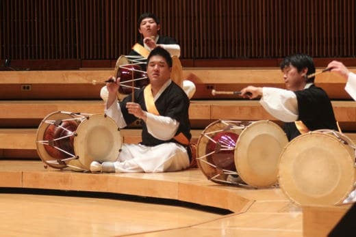 Three men wearing traditional Korean clothing play drums on stage.