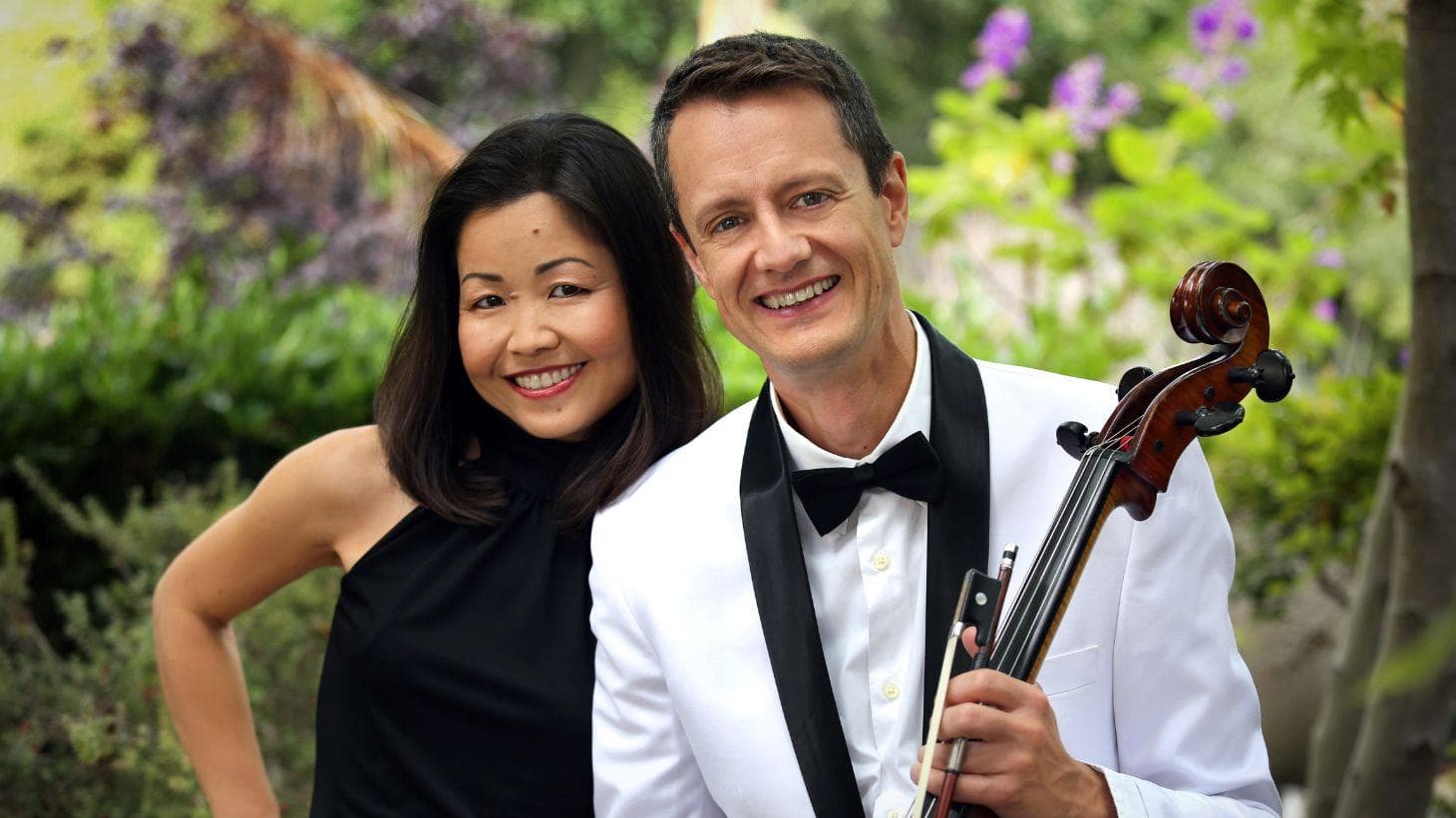 An Asian woman and a white man holding a viola pose for a picture in a garden.