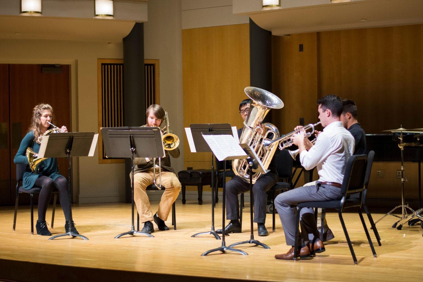Five musicians sit in a semi-circle as they play brass instruments on stage.