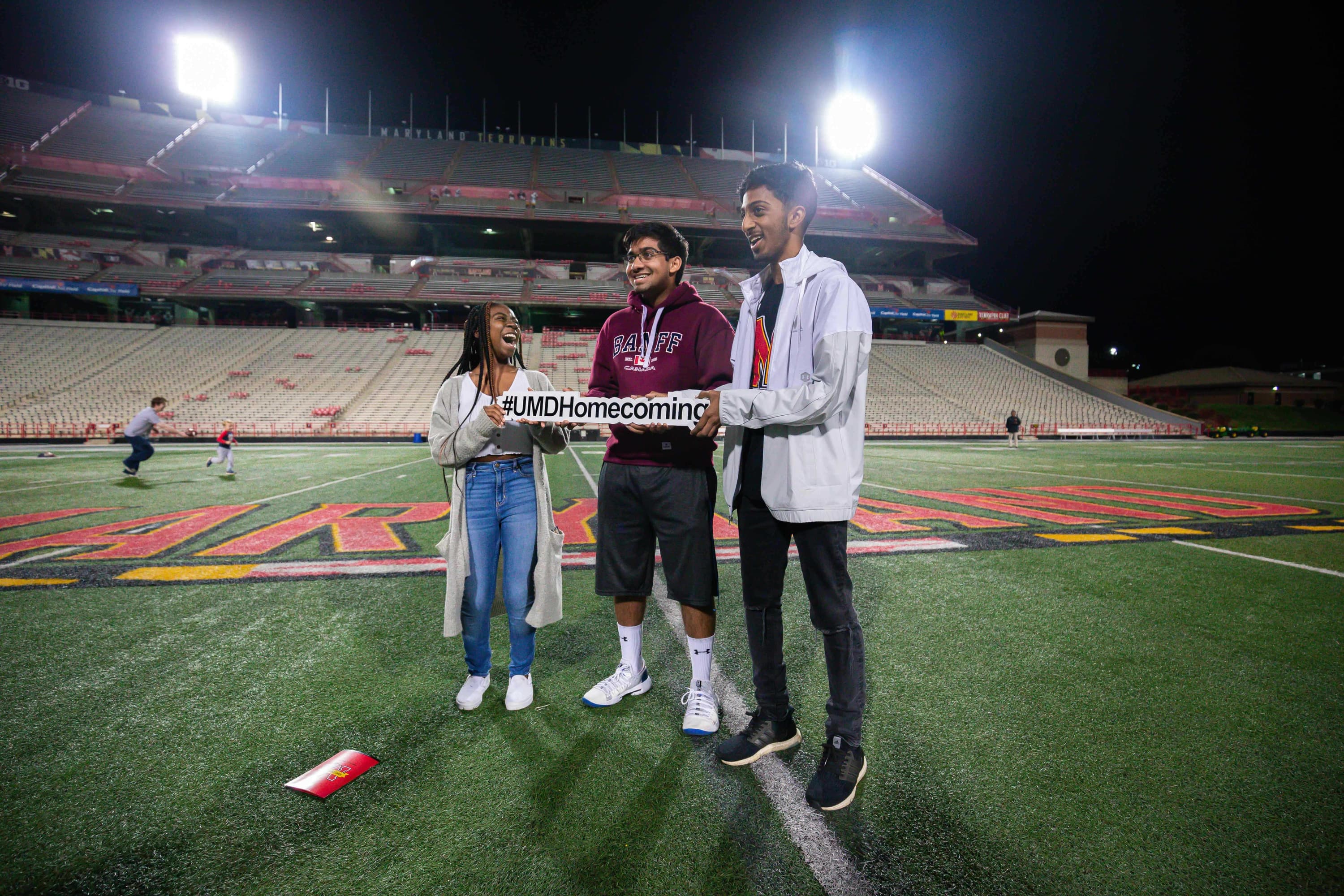 Three students standing on the field in Maryland Stadium holding a sign that says #UMDHomecoming