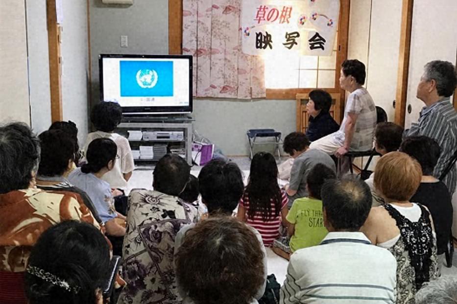 Group of 20 people gather watching a tv screen in a small room.