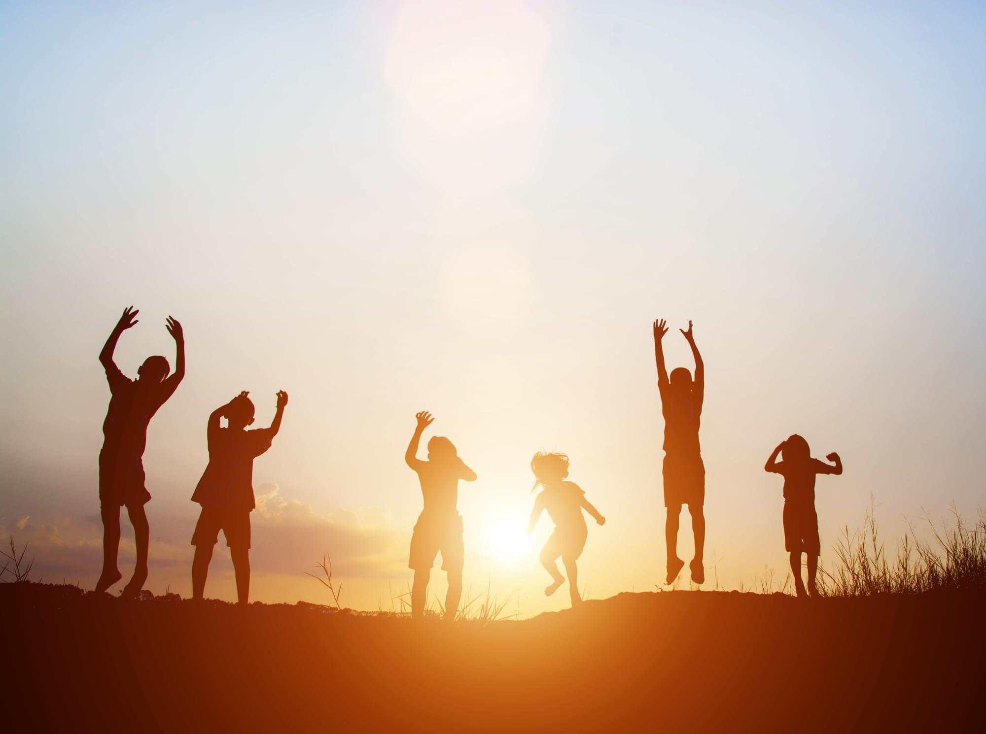 Silhouettes of six children playing outside hands joyfully raised in the air with sunset in background