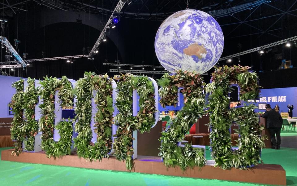 A globe hanging over a sign reading hashtag COP26 with plants growing out of it