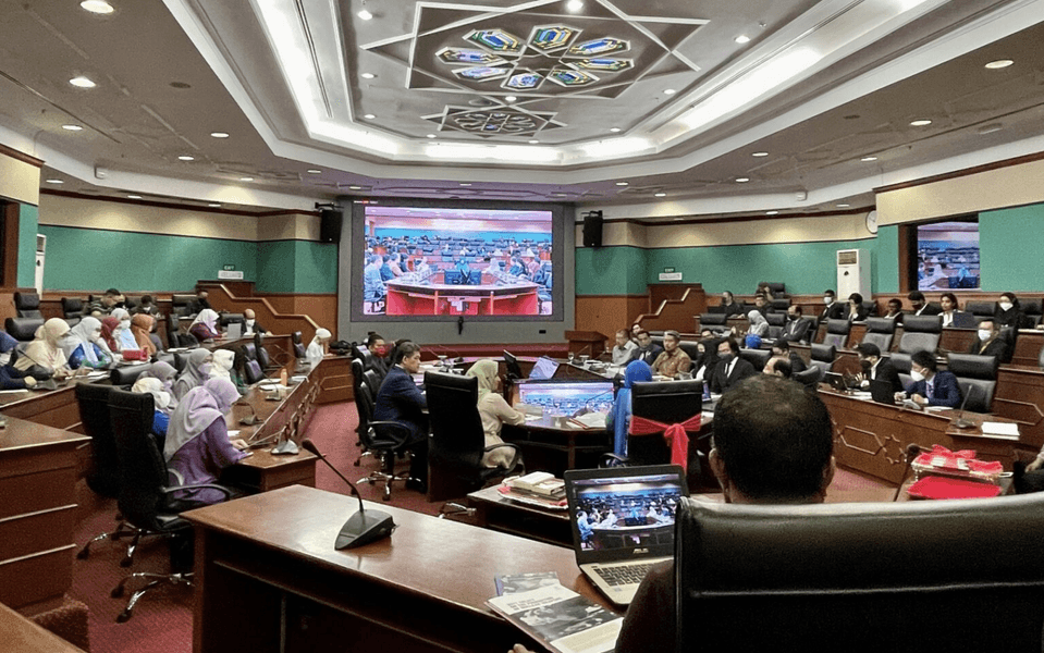 U-shaped conference room filled with people with a screen at the center back