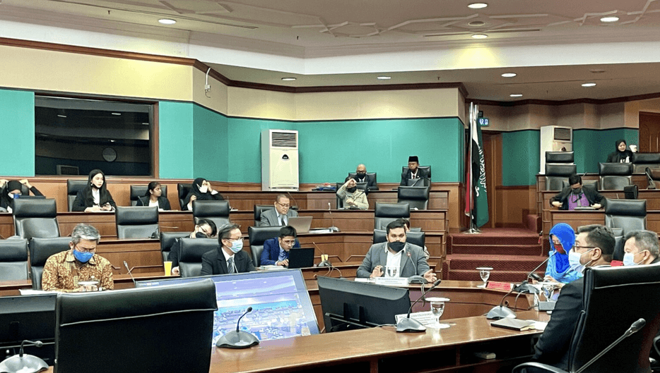 Speaker seated in the bottom center in a conference room filled with people.