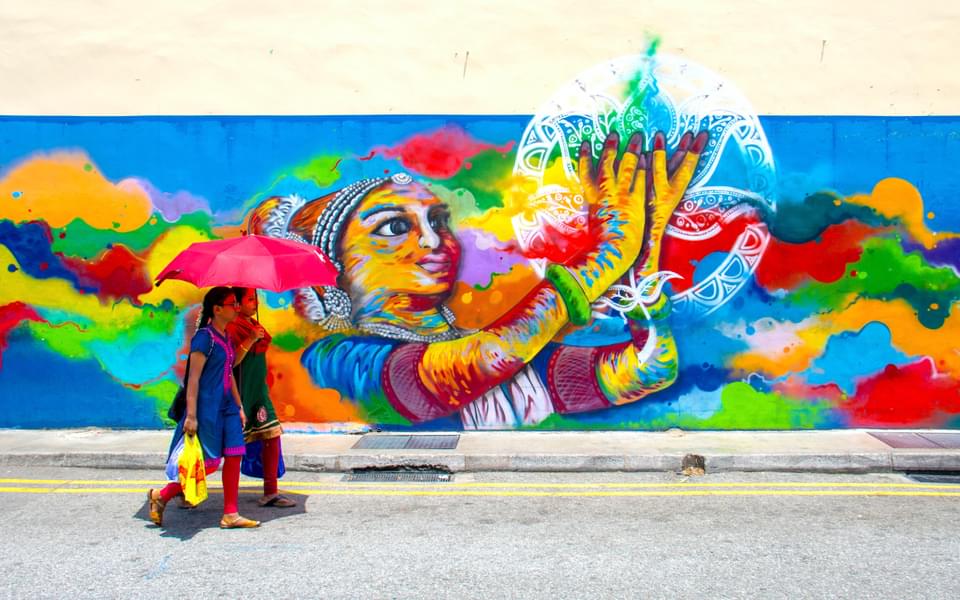 A bright colored mural in the background and two women walking in front of it with an umbrella