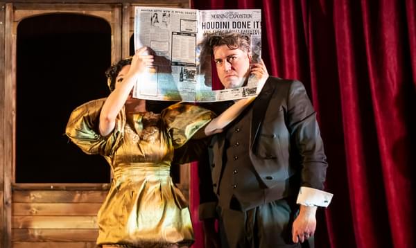 Houdini, dressed in a grey suit, stands next to his glamorous female assistant, dressed in a 1920s-style golden playsuit. She holds up a newspaper with the headline ‘Houdini Done It’. Under the headline, a pane has been cut out of the paper, revealing Houdini’s face where a photo would be.