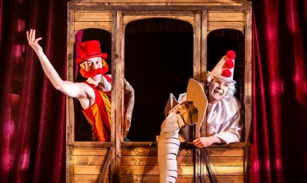 Two actors, one dressed in a red top hat with oversized red handlebar moustache, the other dressed as a clown in a white wig, hat and ruffled top, holding a puppet elephant head. They lean out of the windows of wooden set piece that looks like a train car.