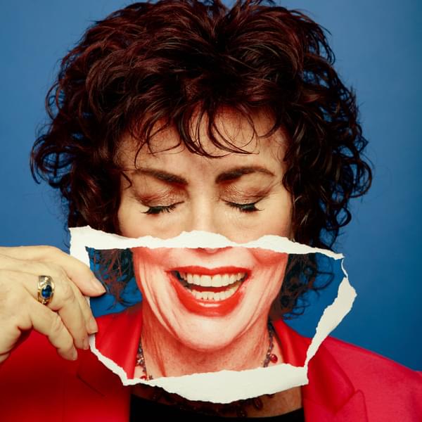 A close up headshot of Ruby Wax on a blue background. She has her eyes closed and is holding a ripped photograph of her mouth in front of her face.