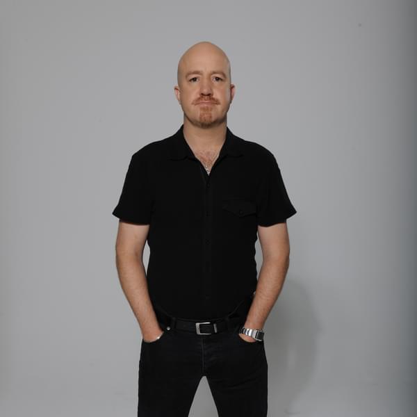 Comedian Andy Parsons. He is Caucasian, bald, wearing a dark shirt with a goatee. The background is grey.