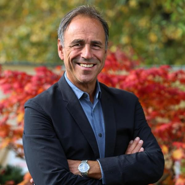 Author Anthony Horowitz smiles into the camera. He is wearing a black suit jacket and blue collared shirt. He has his arms crossed and stands outside in a garden. He is Caucasian, has short grey hair and brown eyes.